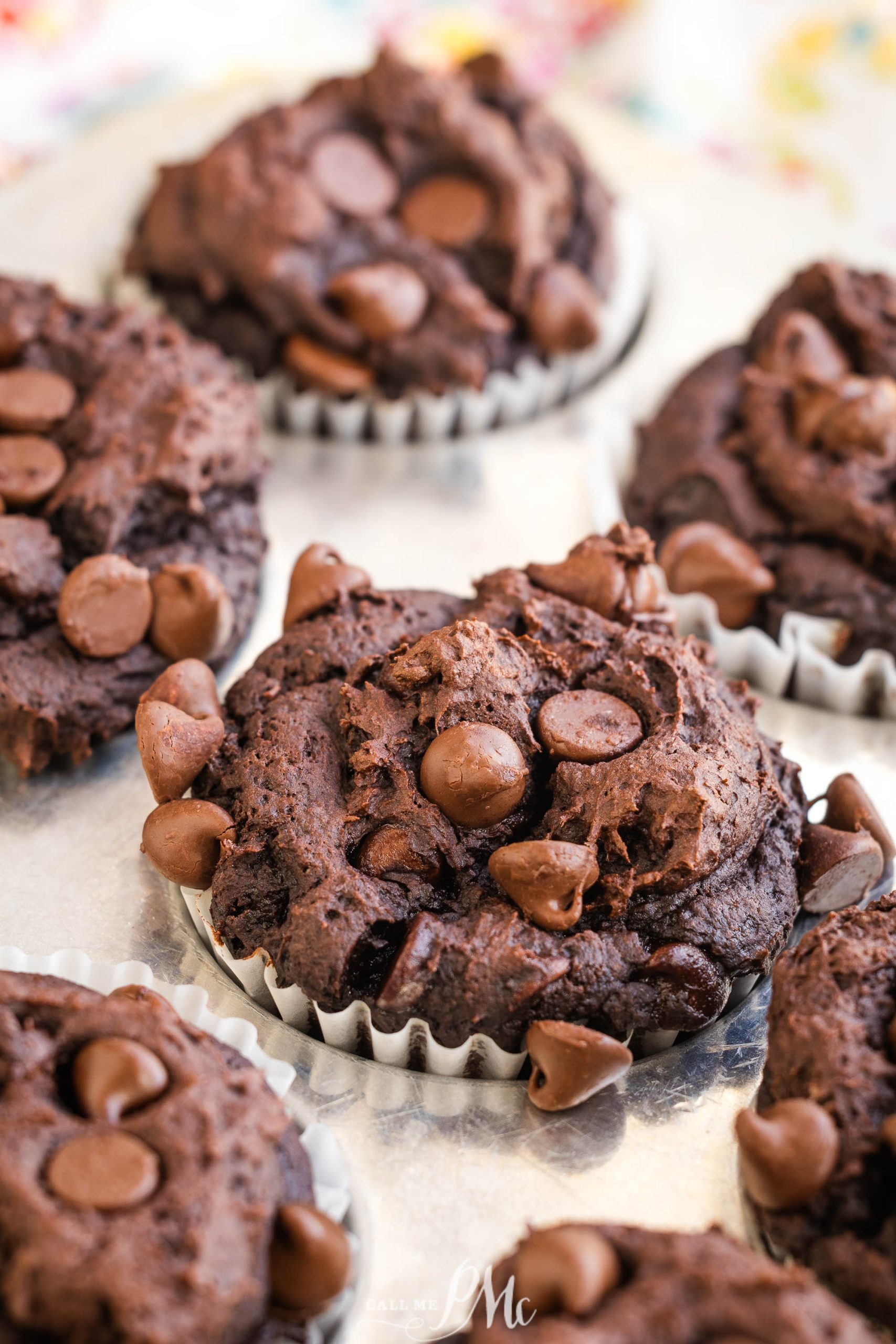 Chocolate muffins with chocolate chips in paper liners arranged on a metallic surface.