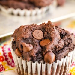 A chocolate muffin with chocolate chips on top, placed in a white paper liner on a colorful floral cloth.