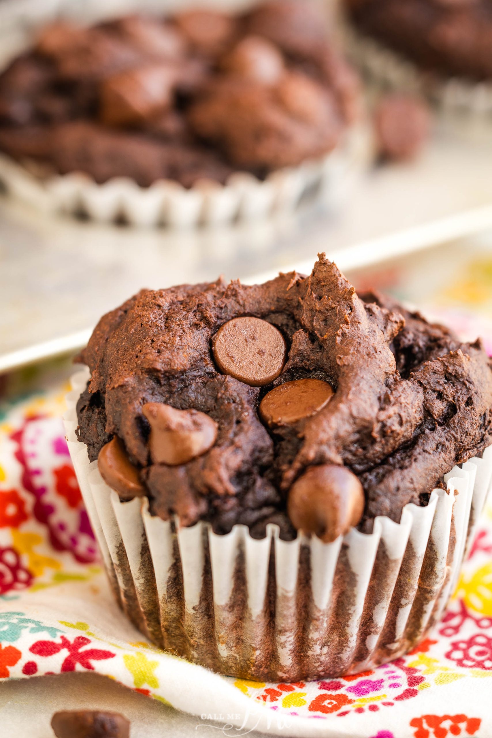 A chocolate muffin with chocolate chips on top, placed in a white paper liner on a colorful floral cloth.