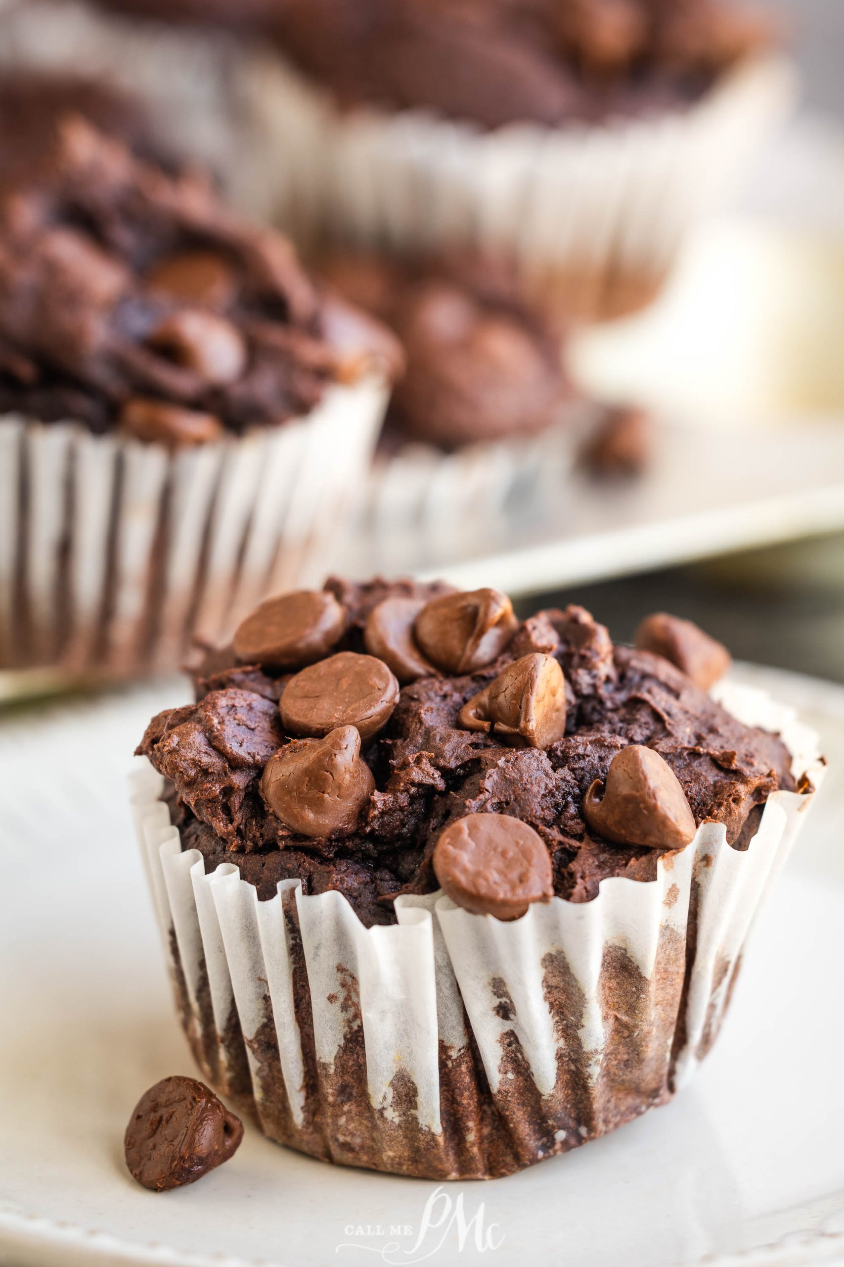 Chocolate muffin with chocolate chips on top, in a white paper wrapper, placed on a decorative plate. More muffins are visible in the background.