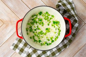 Red pot with chopped onions and green bell peppers on a checkered cloth, set on a wooden surface.