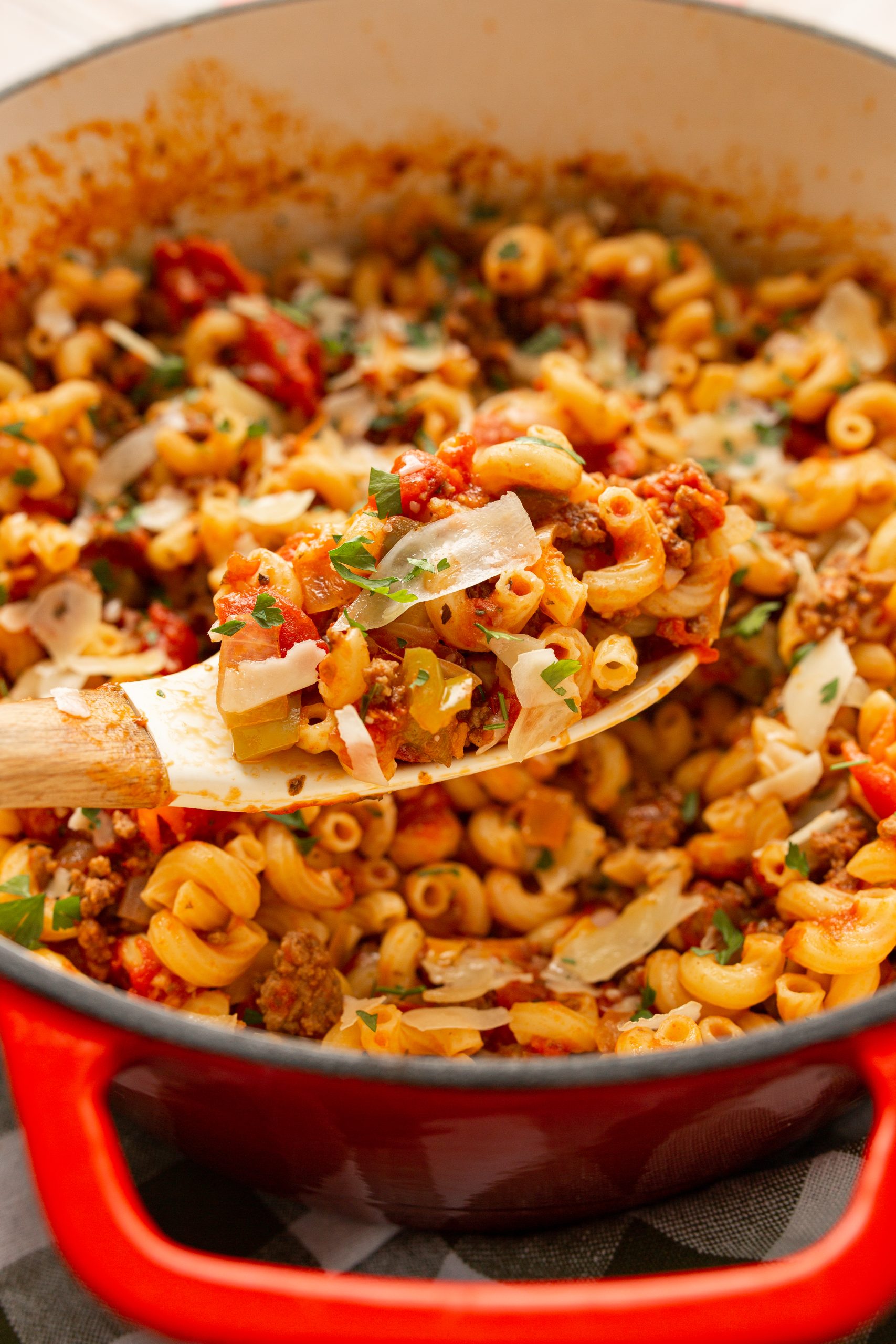 A red pot filled with cooked macaroni in tomato sauce, topped with slices of parmesan and herbs. A wooden spoon lifts a portion of the pasta.