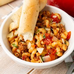 A white bowl of pasta with tomato sauce, ground meat, and shredded cheese, topped with two breadsticks. A red pot and a single breadstick are in the background.