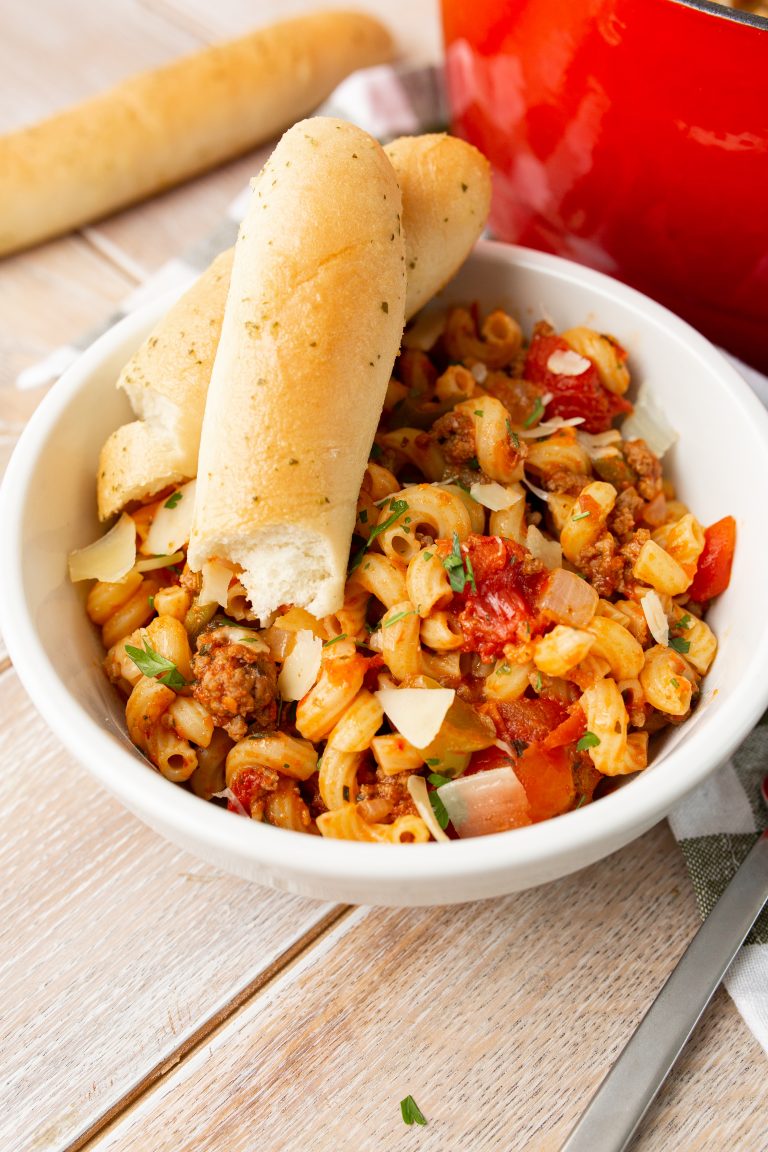 A white bowl of pasta with tomato sauce, ground meat, and shredded cheese, topped with two breadsticks. A red pot and a single breadstick are in the background.