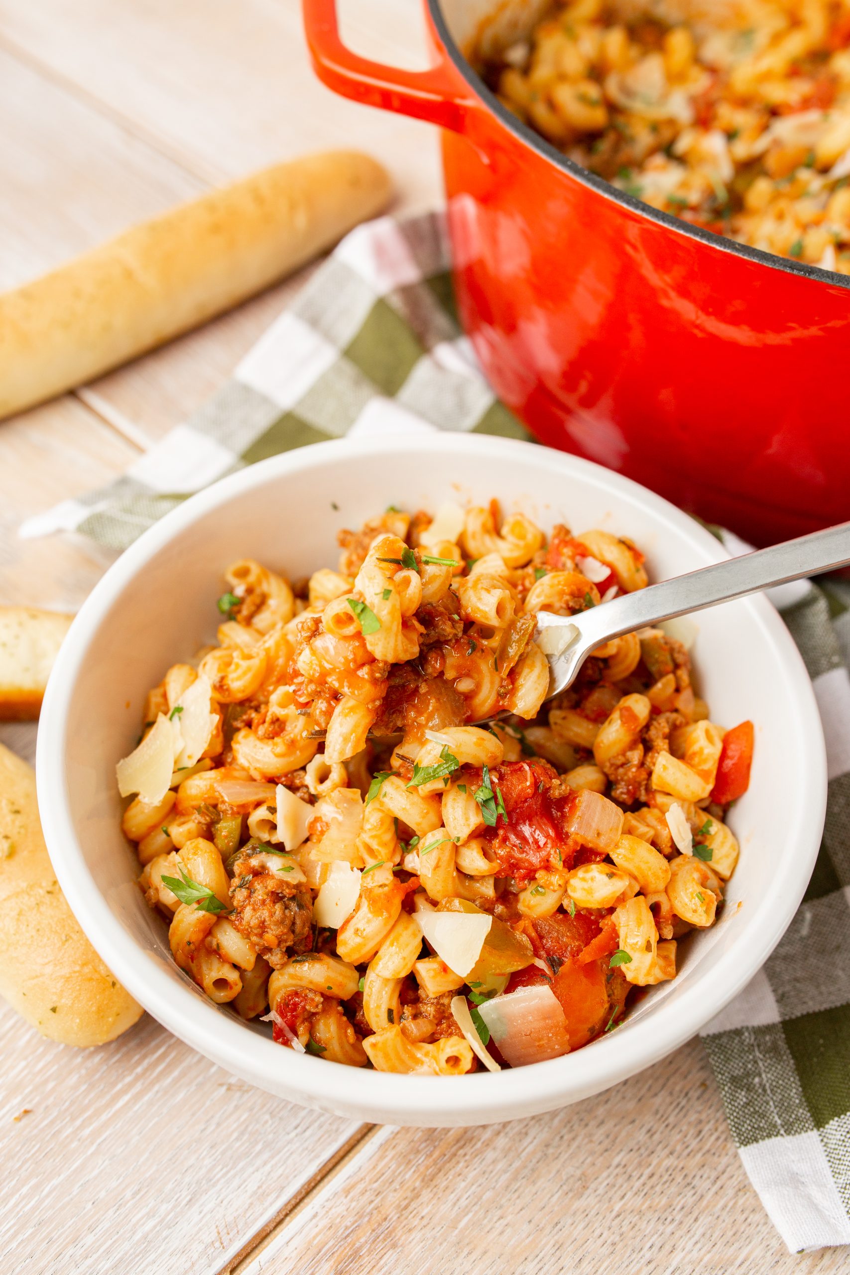 A bowl of American chop suey, topped with herbs, next to a red pot and breadsticks on a checkered cloth.