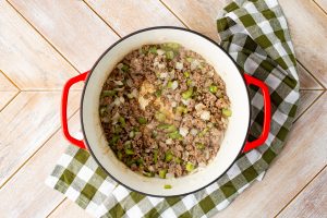 Red pot with ground meat, diced onions, and green peppers on a checkered cloth.