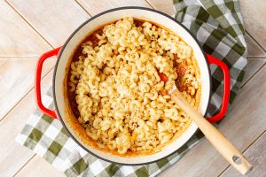 A red pot filled with cooked pasta sits on a checkered cloth. A wooden spoon rests inside the pot.