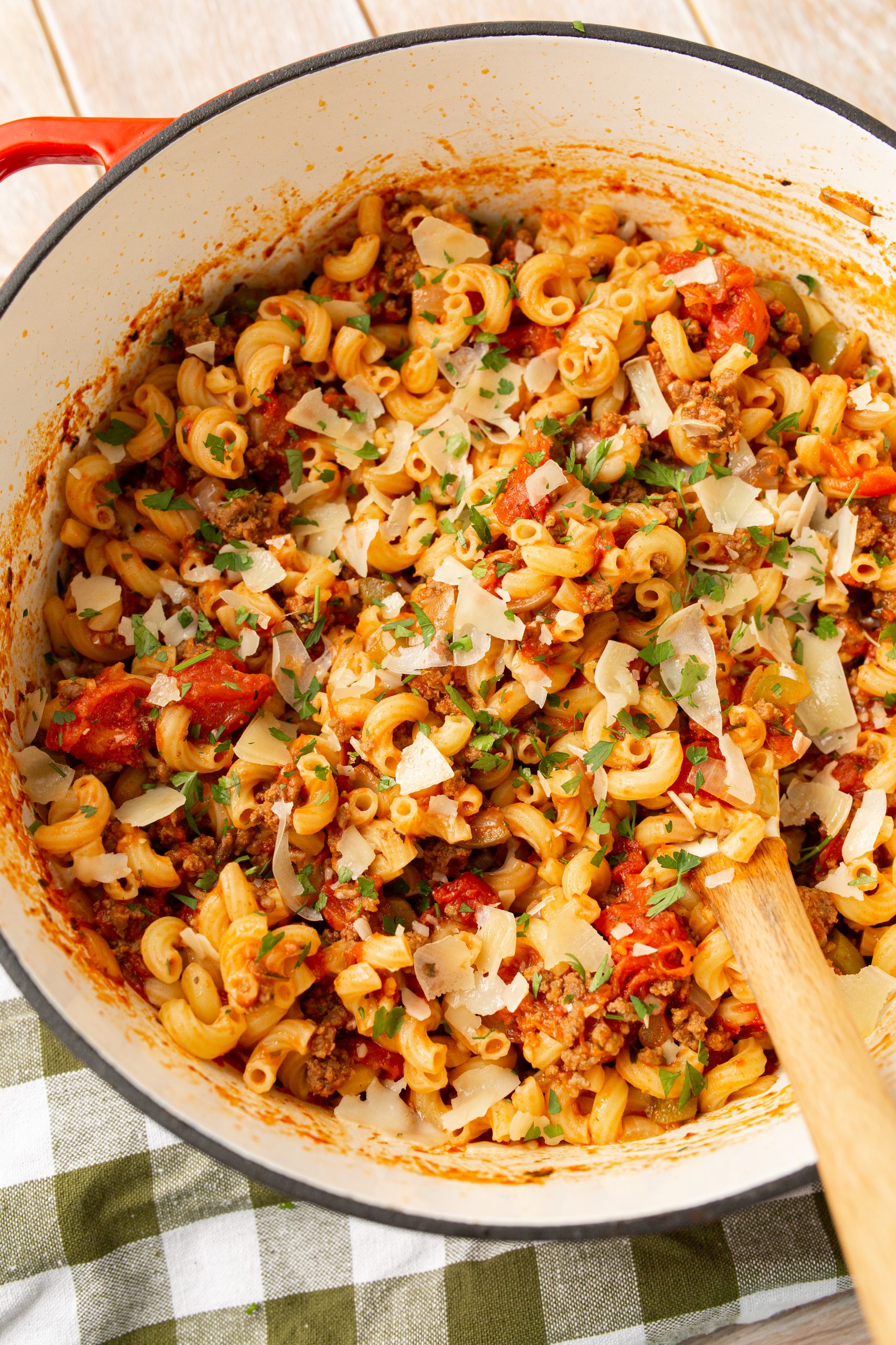 A pot of American chop suey and garnished with chopped herbs and grated cheese, with a wooden spoon on the side.