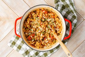 A pot of cooked macaroni mixed with vegetables, herbs, and shaved cheese on a red-handled pot, placed on a green checkered cloth.