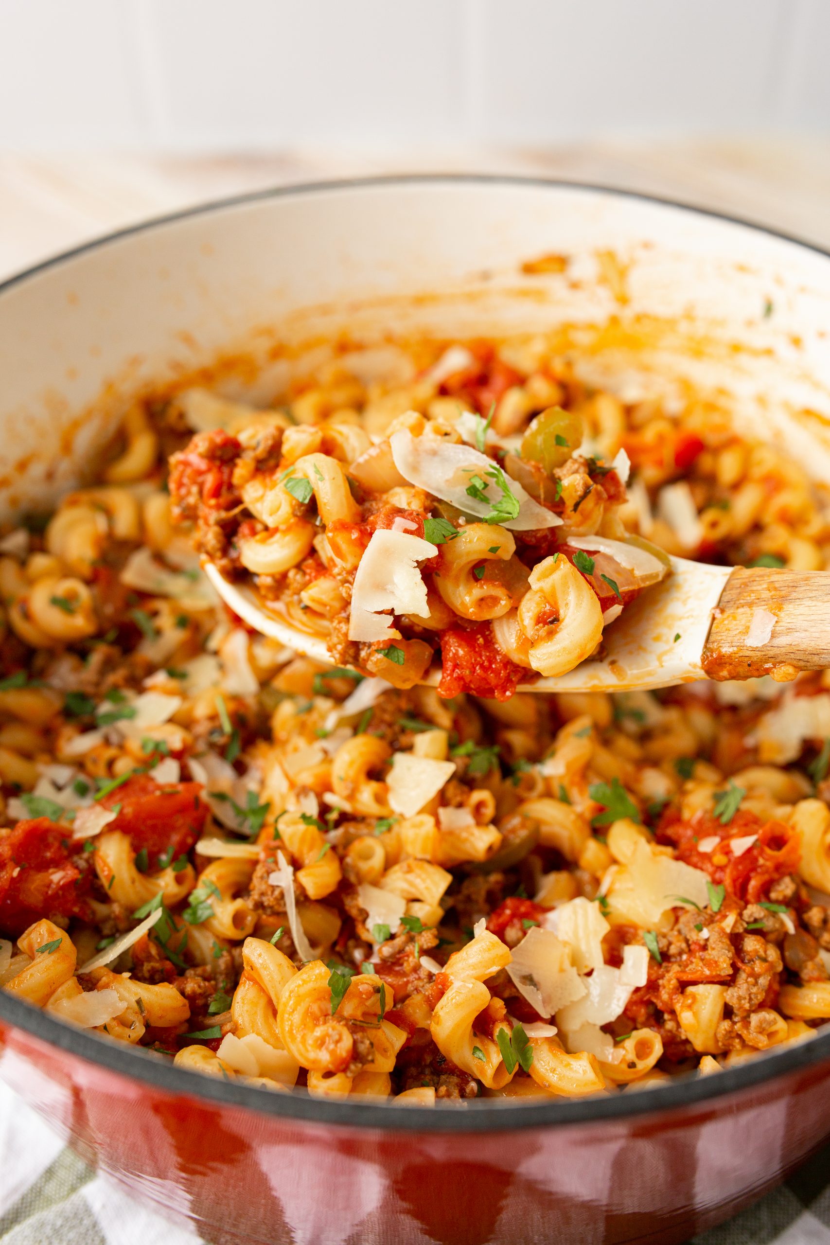 A pot of elbow pasta with a rich tomato and meat sauce, garnished with herbs and cheese, shown with a wooden spoon lifting a portion.