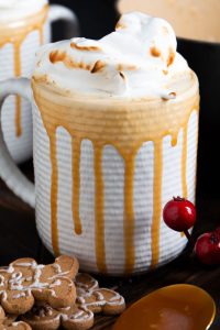 A mug filled with creamy brown butter eggnog topped with whipped cream and caramel drizzle, accompanied by gingerbread cookies and red berries.