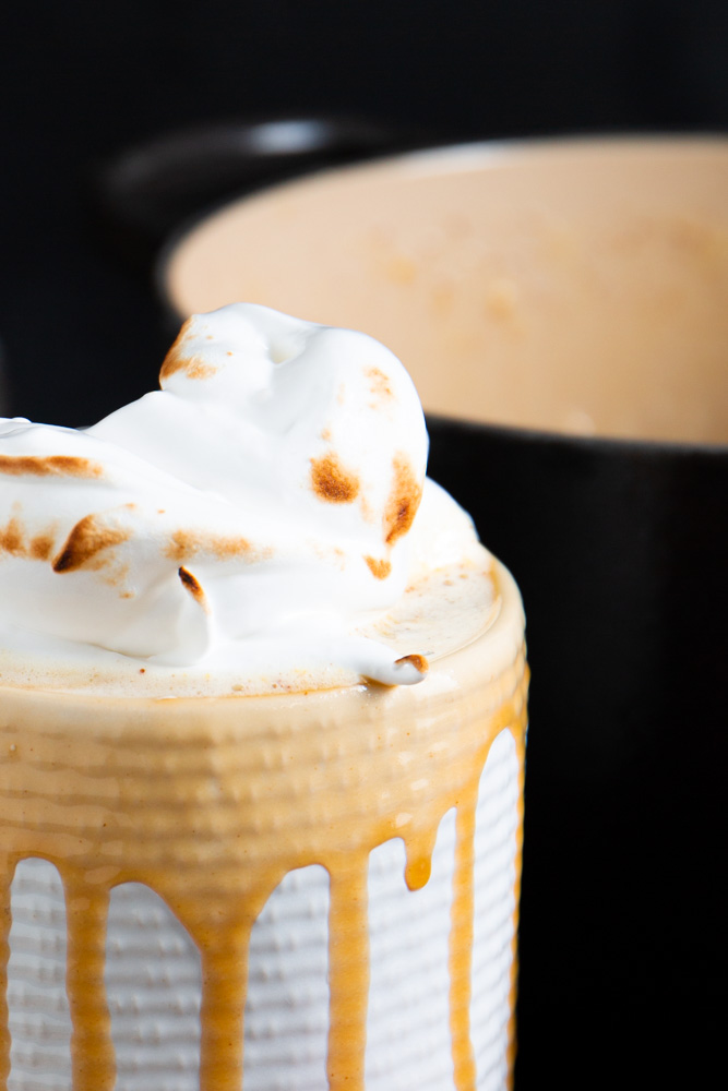 A close-up of a mug with hot chocolate, topped with toasted whipped cream and a hint of brown butter eggnog. Drips of chocolate run down the mug's sides, adding to the rich indulgence.