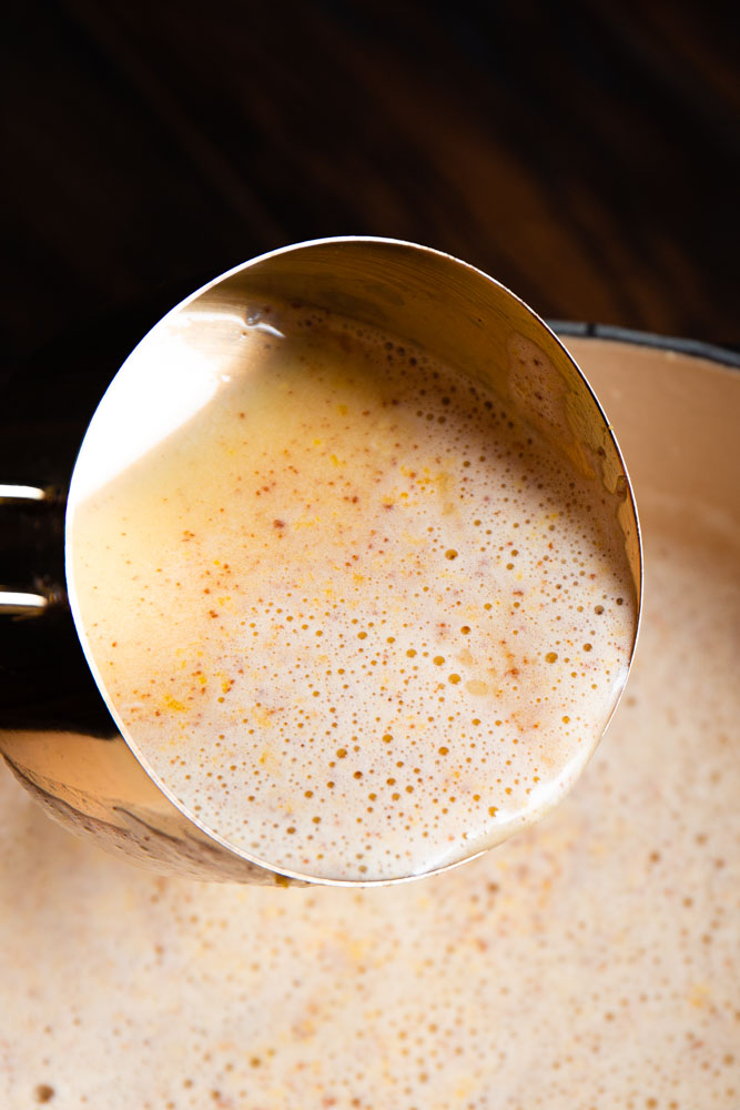 Close-up of a ladle scooping frothy brown butter eggnog speckled with spices from a pot.