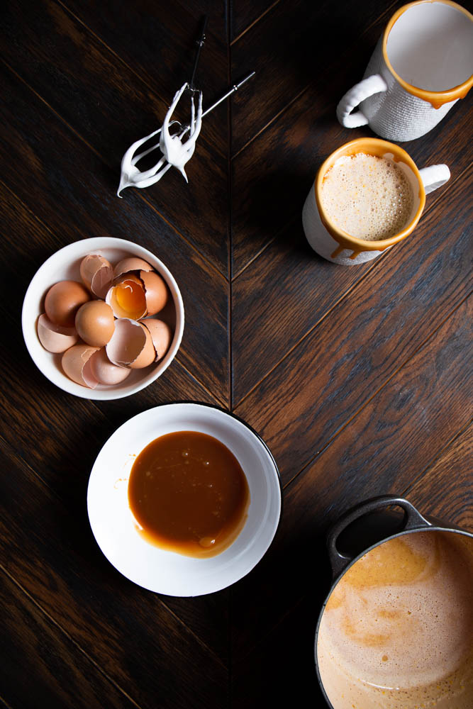 A wooden table set with a bowl of egg shells, a mug, a bowl, a saucepan, and a whisk attachment coated in cream.