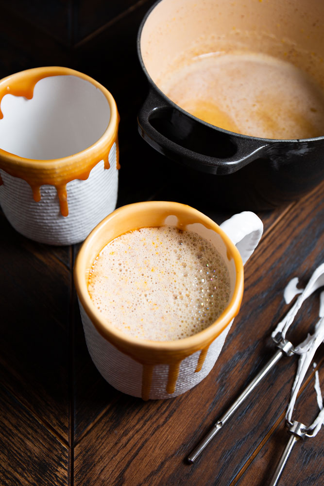 Two white ceramic mugs with caramel drips are filled with frothy, brown butter eggnog. A black pot containing more of the creamy concoction is nearby, along with two metal stirring utensils.