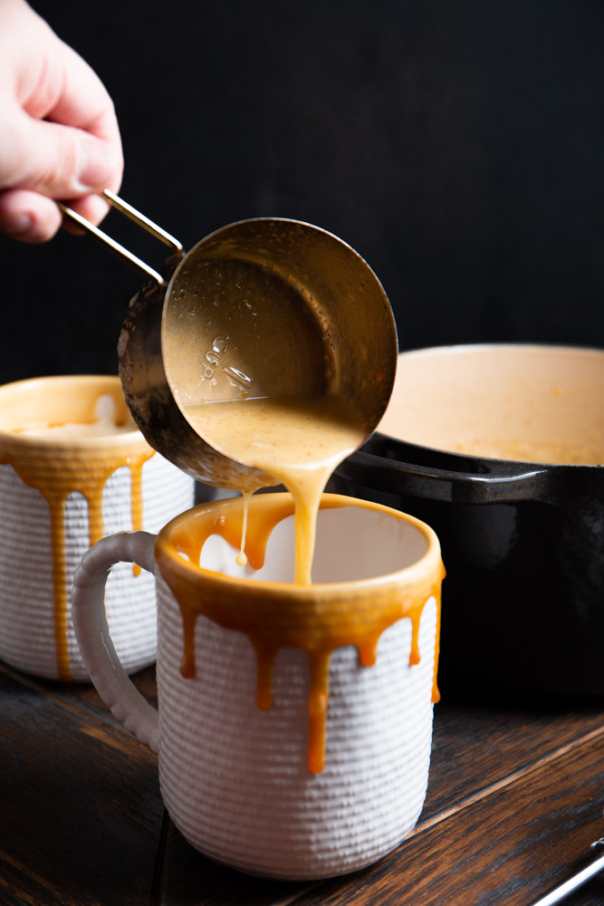 A hand pours creamy soup from a measuring cup into a mug with a decorative dripped glaze, reminiscent of the rich hues of brown butter. Additional mugs and a black pot rest in the background on the wooden surface, evoking cozy gatherings and winter treats like homemade eggnog.