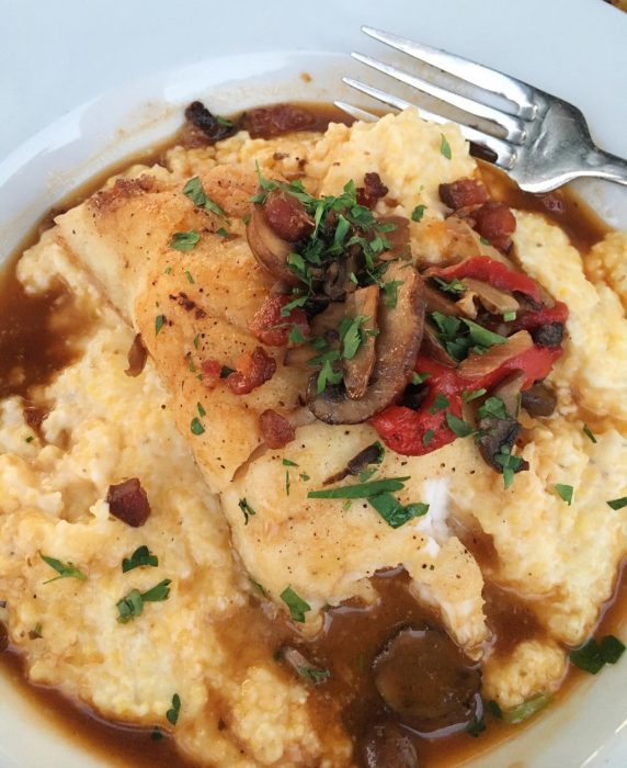 A dish of fish on a bed of creamy yellow grits, topped with mushrooms, red peppers, and herbs, with a fork on the side.