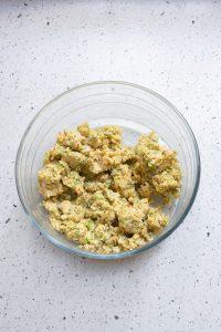 A clear bowl containing a mixture of chopped vegetables and grains, accompanied by a side of Cheesy Broccoli Bites, rests on a speckled countertop.