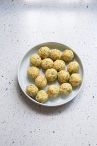 A plate with 14 round, uncooked falafel balls rests on a speckled countertop, reminiscent of cheesy broccoli bites in their perfect symmetry.