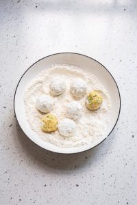 A white plate with flour-covered round dough balls, some stuffed with cheesy broccoli goodness, sits alongside a few uncovered rounds on a speckled countertop.