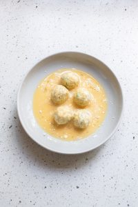 Five dough balls, soon-to-be Cheesy Broccoli Bites, rest in a bowl of beaten eggs on a speckled countertop.