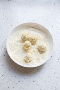 Six round balls covered in breadcrumbs rest on a white plate, placed on a speckled countertop. These delightful treats are actually Cheesy Broccoli Bites, offering a tasty twist with every crunchy mouthful.