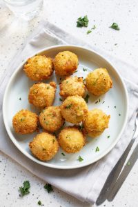 A plate of fried arancini balls, reminiscent of cheesy broccoli bites, is garnished with chopped parsley on a white cloth. A fork and knife are placed nearby on the table.