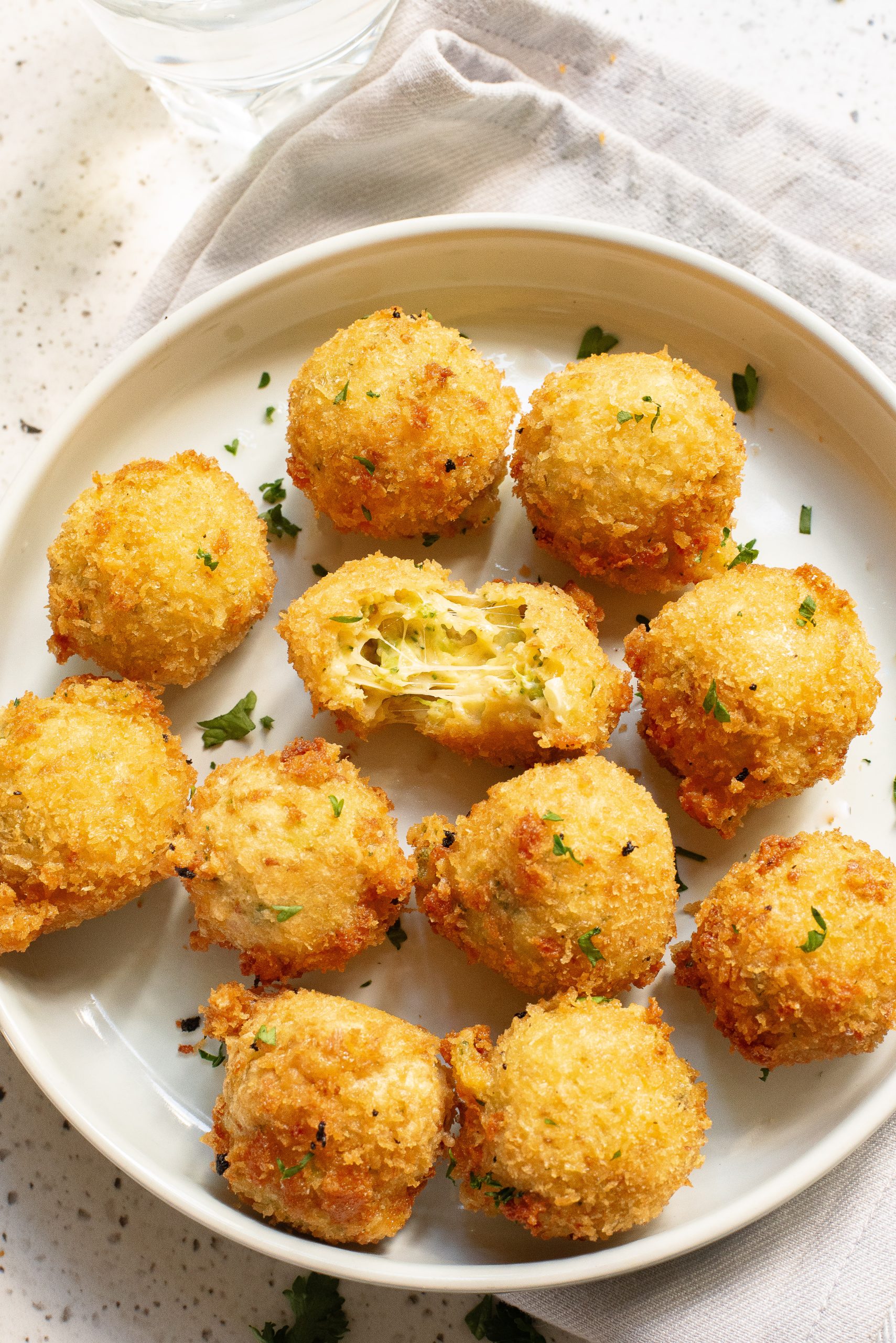 A white plate with nine golden-brown, fried balls. One ball is split open, revealing a soft, yellow filling. Garnished with chopped herbs.