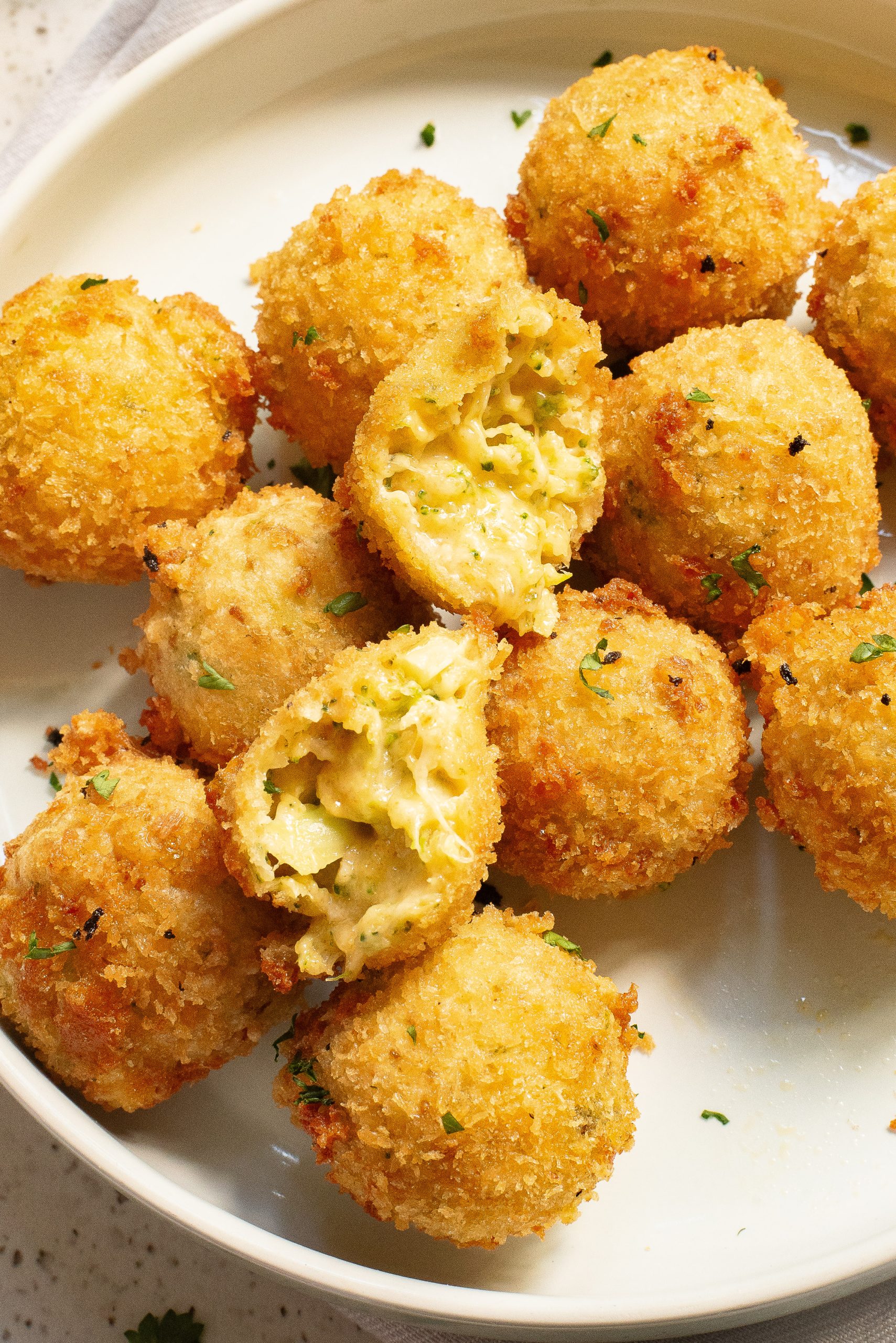 A plate of golden-brown Cheesy Broccoli Bitesgarnished with herbs. One ball is cut open, revealing the creamy pasta filling inside.