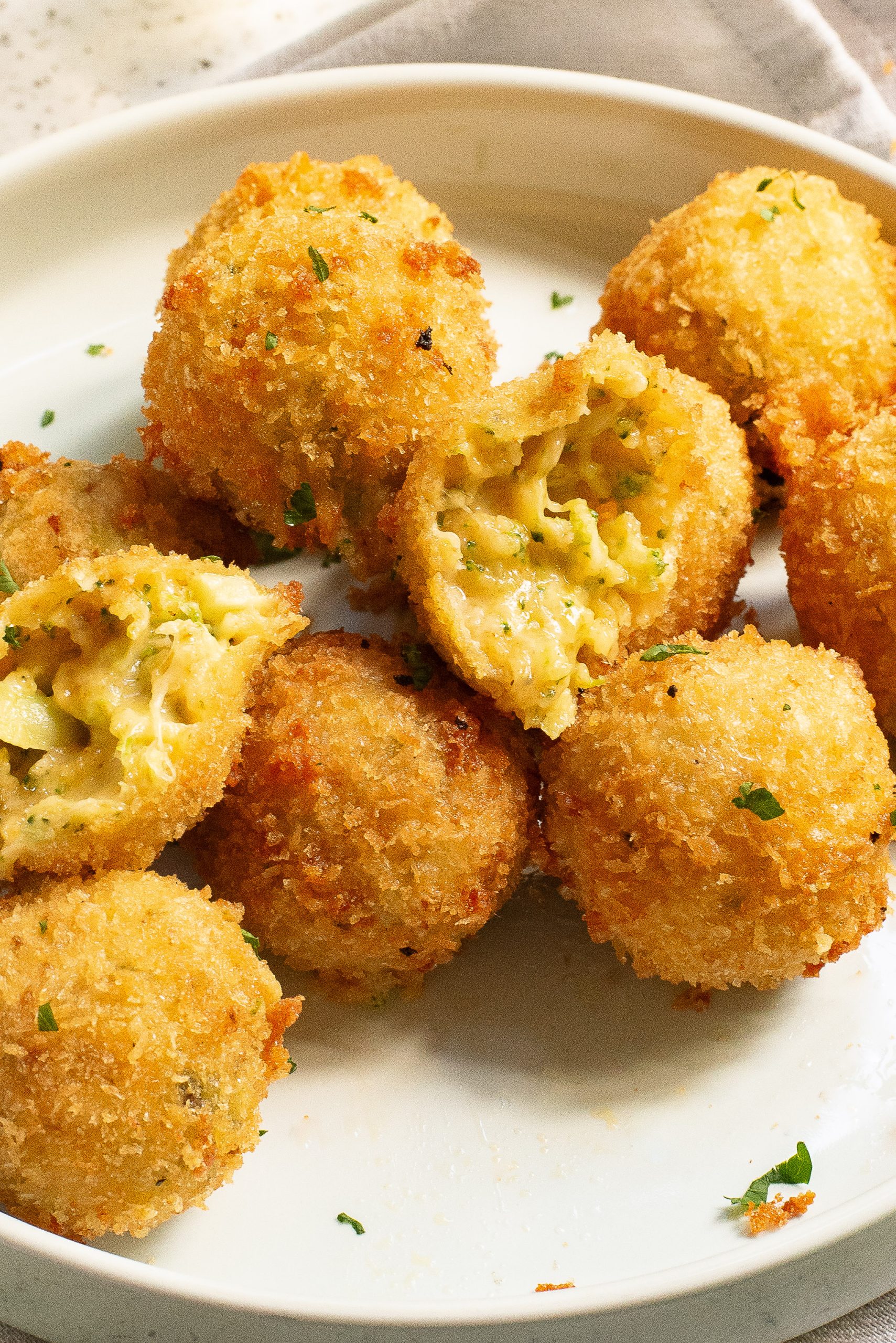 A plate of golden-brown, fried cheesy broccoli bites displayed, with one ball broken open to reveal the creamy pasta and broccoli filling inside.