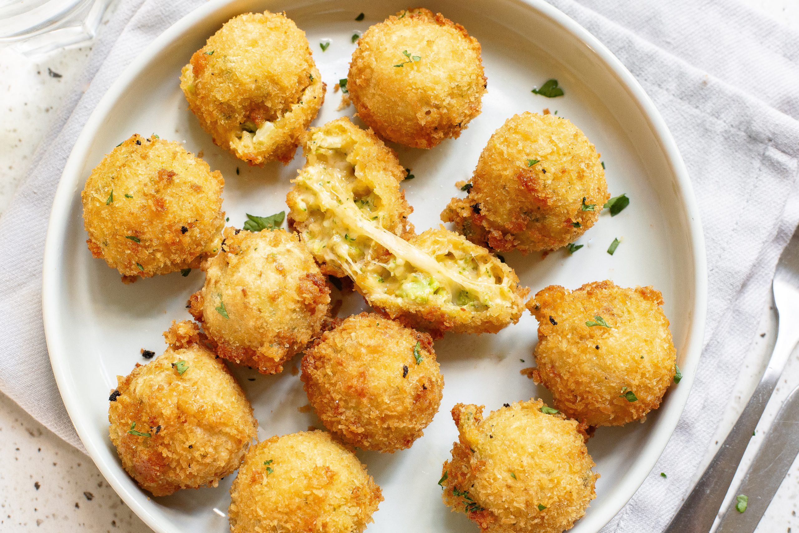 A white plate with nine breaded Cheesy Broccoli Bites, one halved revealing a cheesy filling with green bits, possibly herbs or vegetables, on a white cloth.