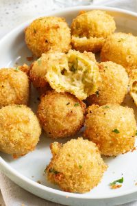 A white plate with golden-brown, crispy arancini balls; one is cut open, revealing a creamy, cheesy filling with herbs.