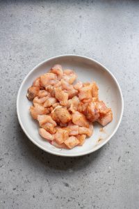 Diced raw chicken pieces seasoned with spices in a white bowl placed on a speckled gray countertop.