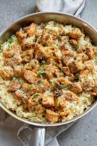 A skillet filled with seasoned chicken pieces over a bed of rice, garnished with herbs.