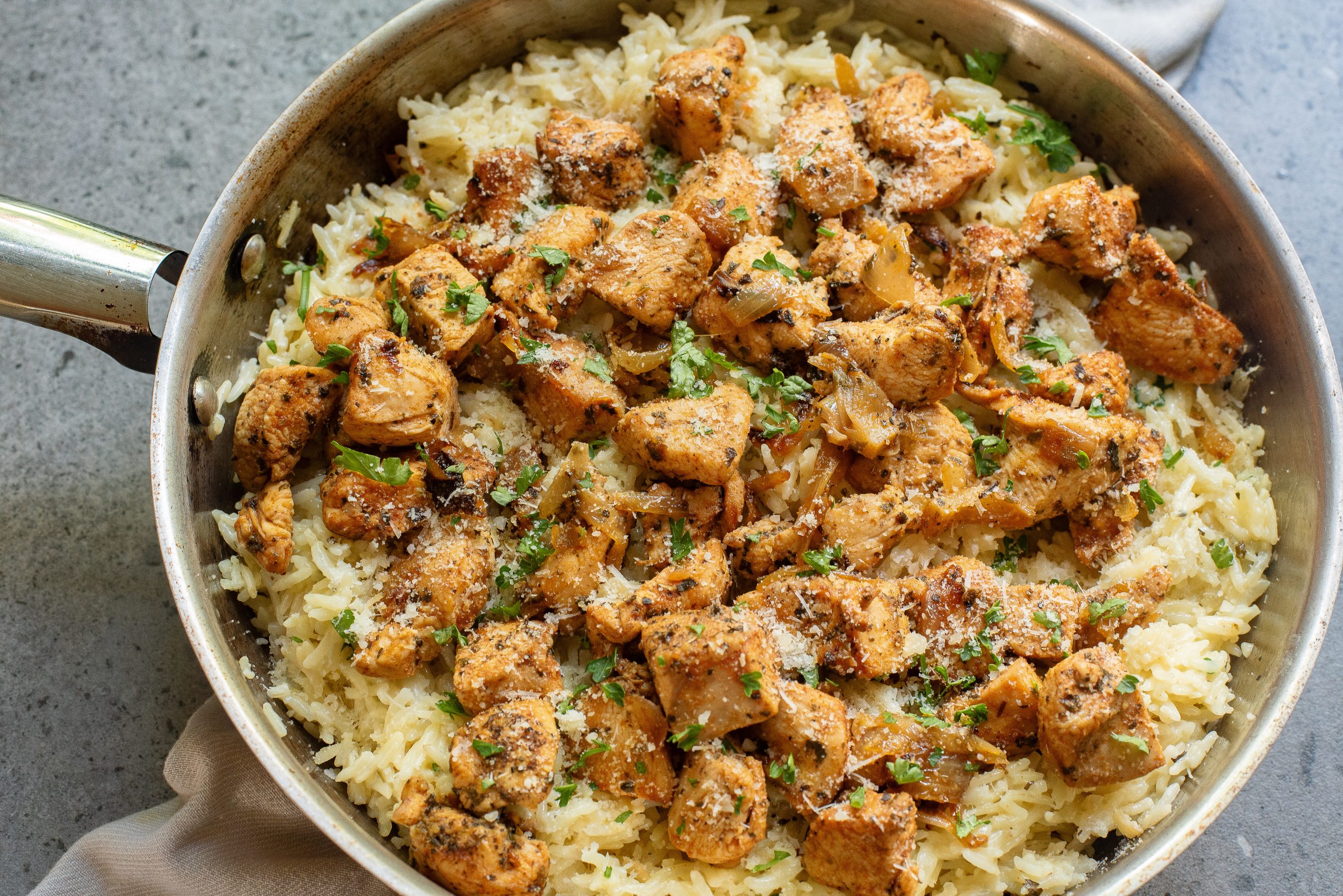 Pan with seasoned chicken pieces on a bed of rice garnished with chopped herbs and grated cheese.