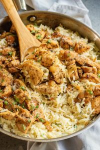 A skillet filled with seasoned chicken pieces and rice, garnished with herbs, with a wooden spoon resting on top.