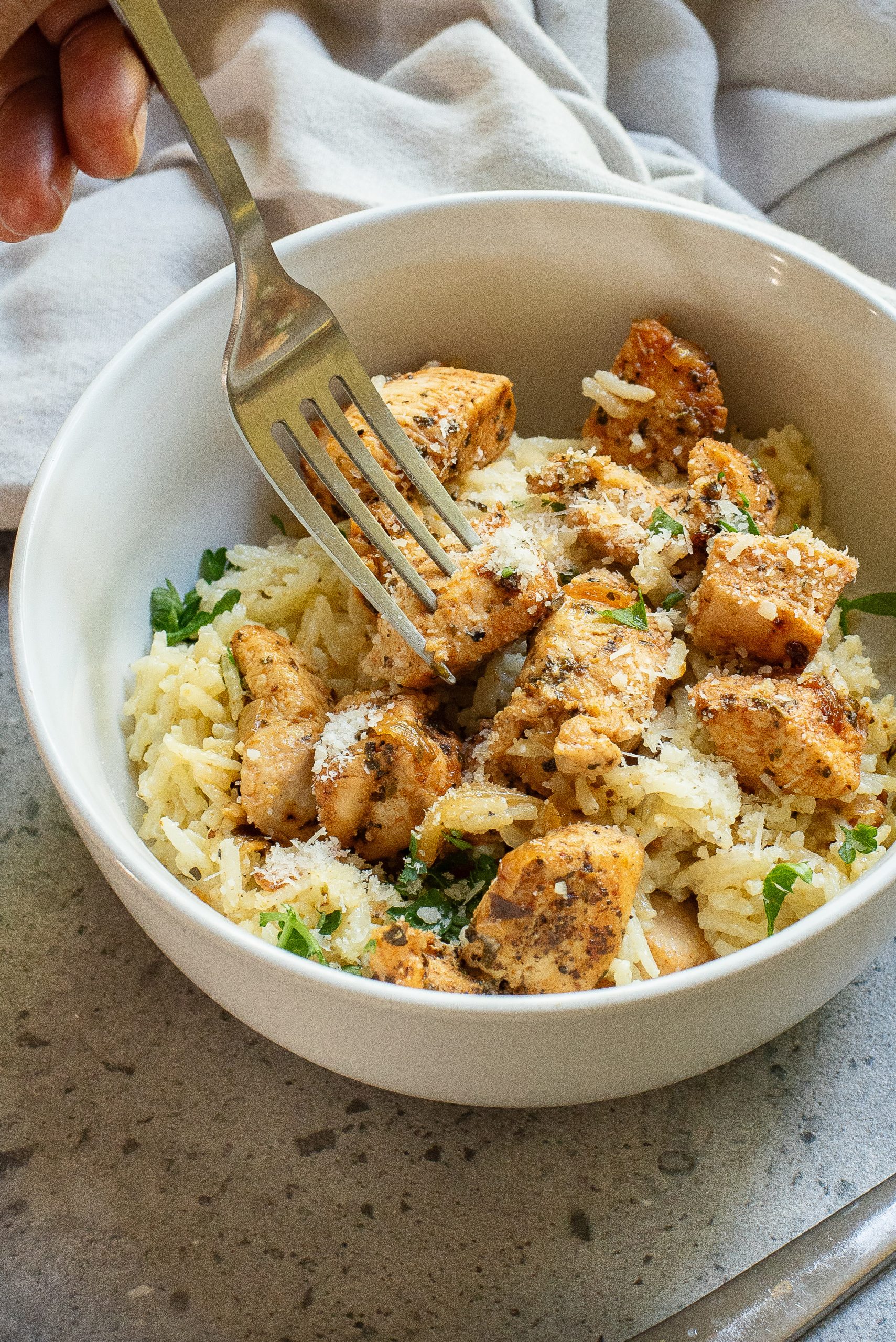 A fork poised over a bowl of seasoned chicken pieces on a bed of rice, garnished with herbs and grated cheese.