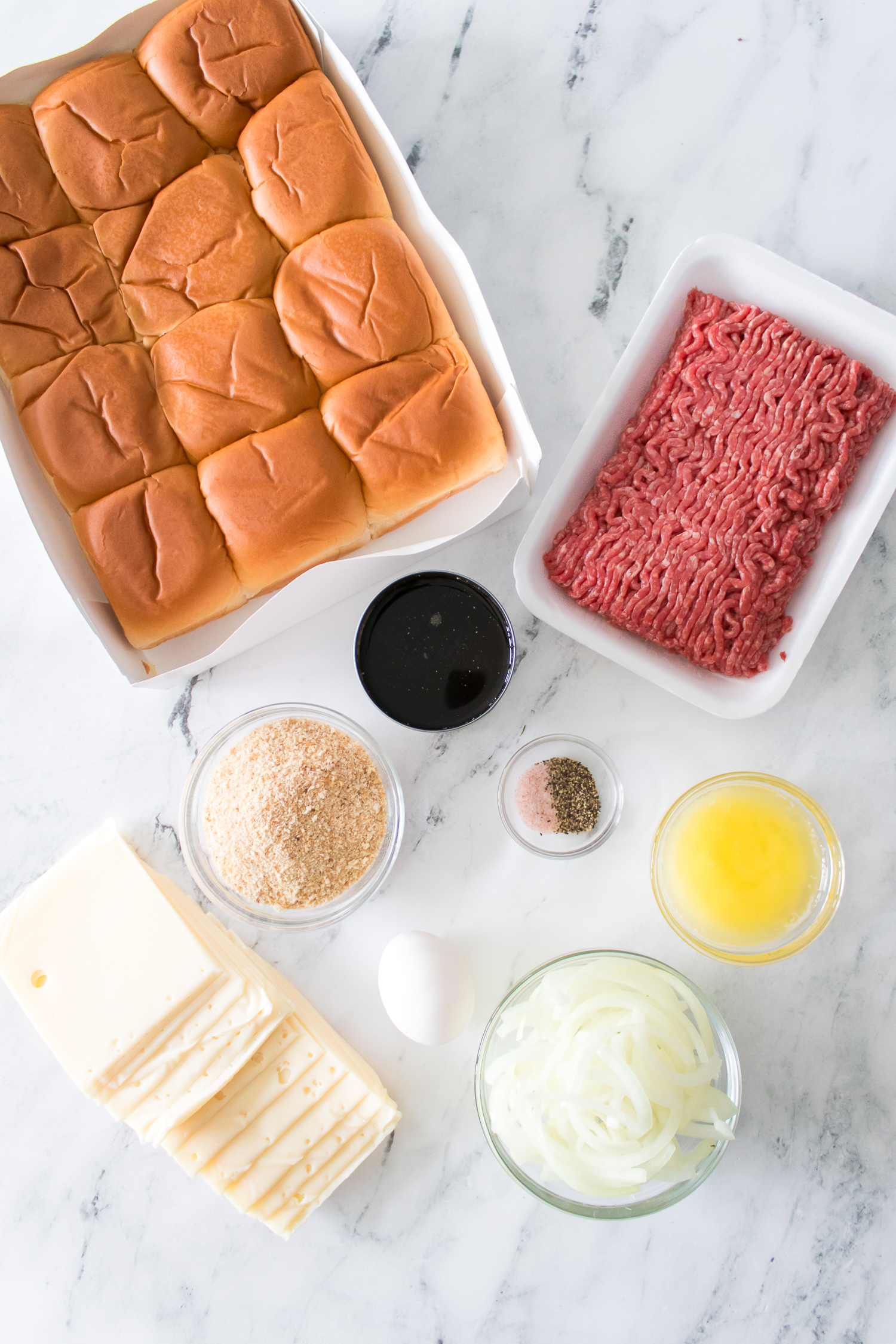 Ingredients for sliders on a marble surface: a tray of rolls, ground beef, Worcestershire sauce, breadcrumbs, sliced cheese, egg, diced onion, seasoning, and melted butter.