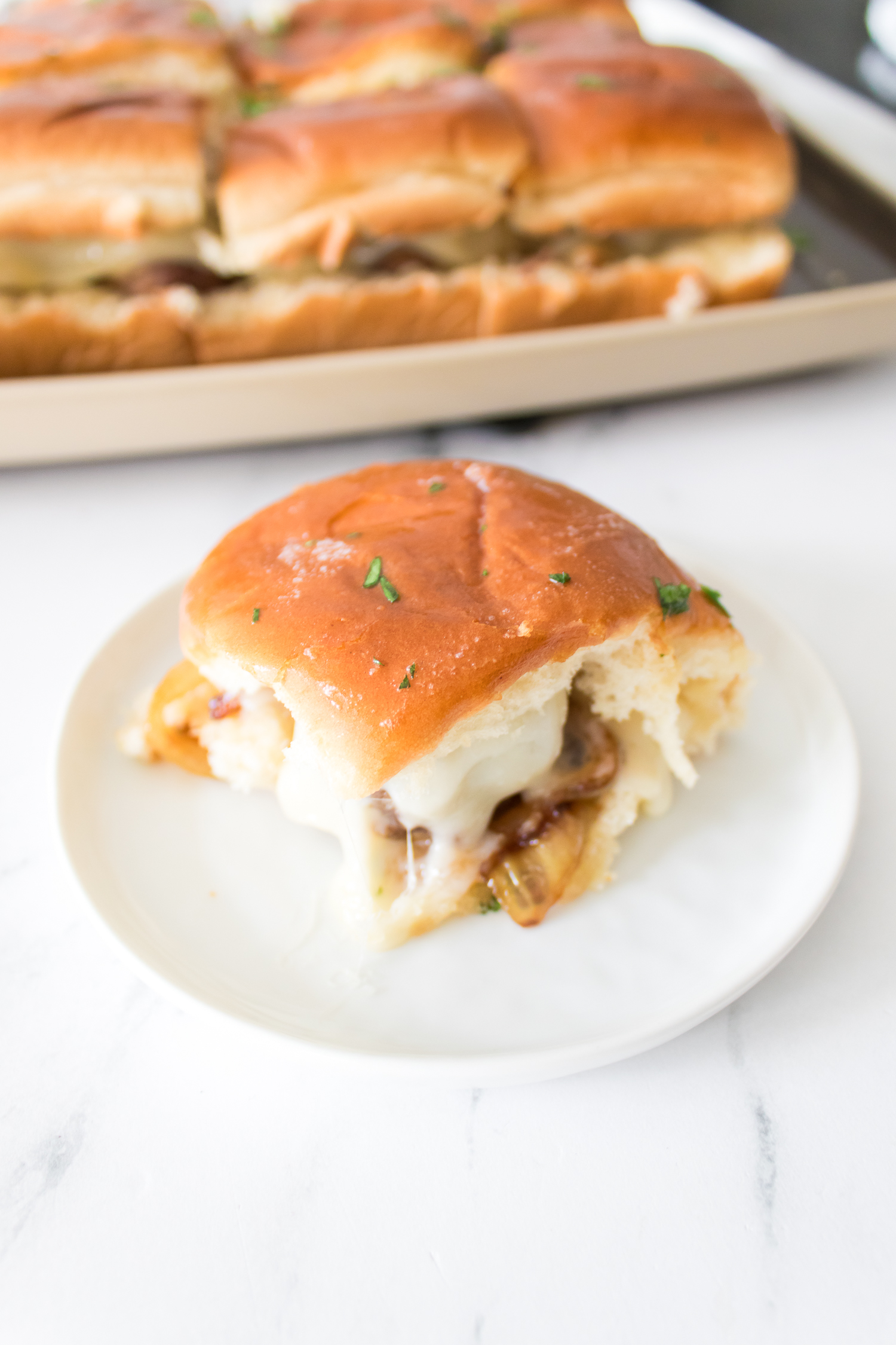 A sandwich with melted cheese and caramelized onions on a small plate, with more sandwiches on a tray in the background.