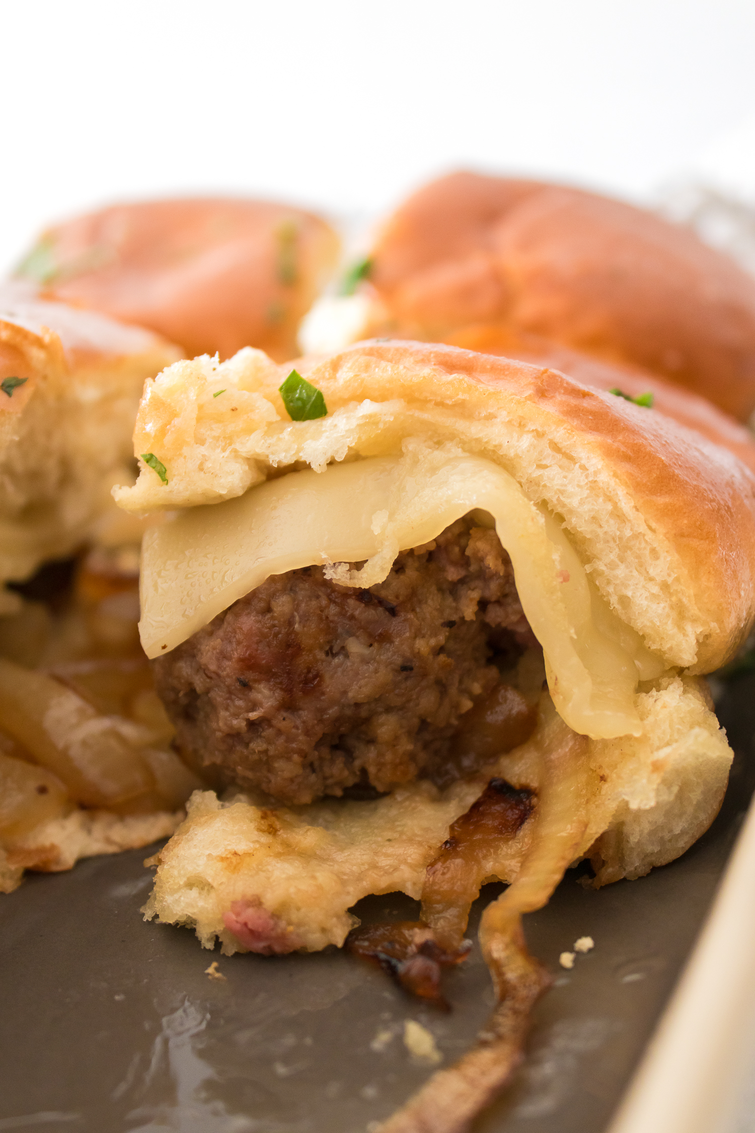 Close-up of a beef slider with melted cheese and caramelized onions on a soft bun. Two more sliders are partly visible in the background.