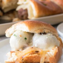 Close-up of a meatball slider with melted cheese and sautéed onions on a small white plate. A tray of additional sliders is in the background.