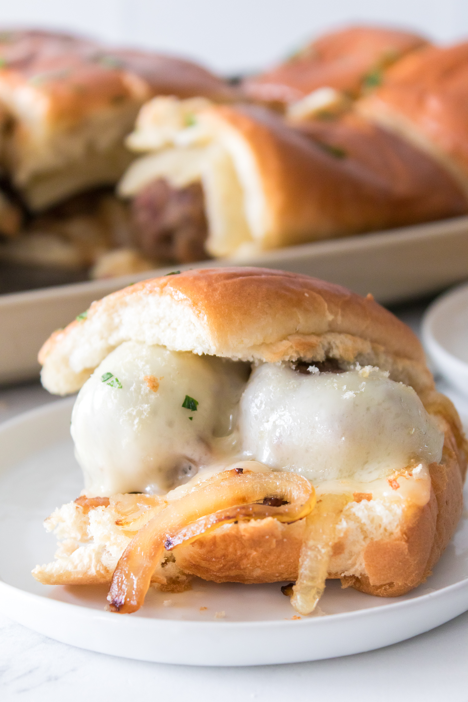 Close-up of a meatball slider with melted cheese and sautéed onions on a small white plate. A tray of additional sliders is in the background.