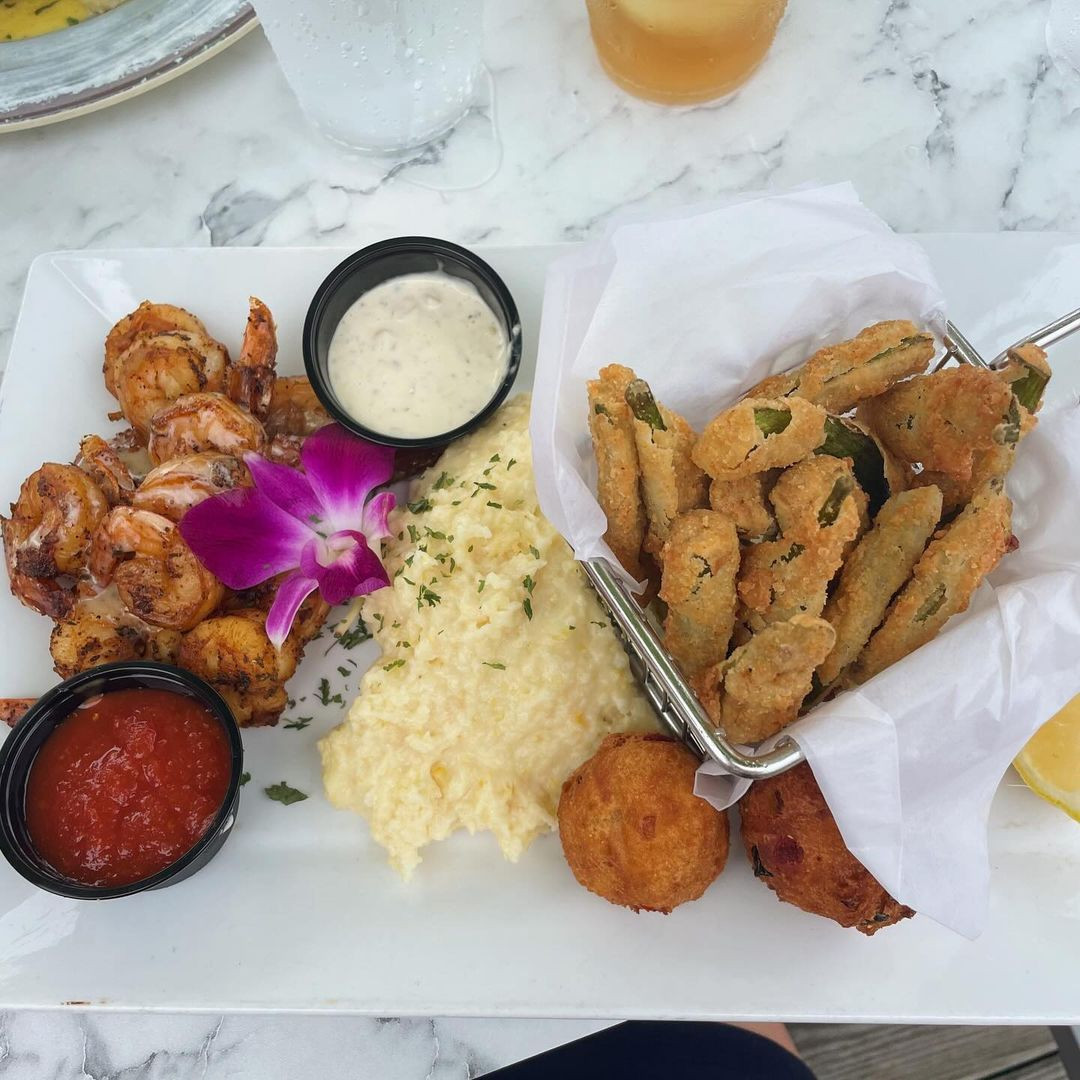 A plate with grilled shrimp, grits, fried okra, two hush puppies, two dipping sauces, and a garnish of purple orchid on a marble table.