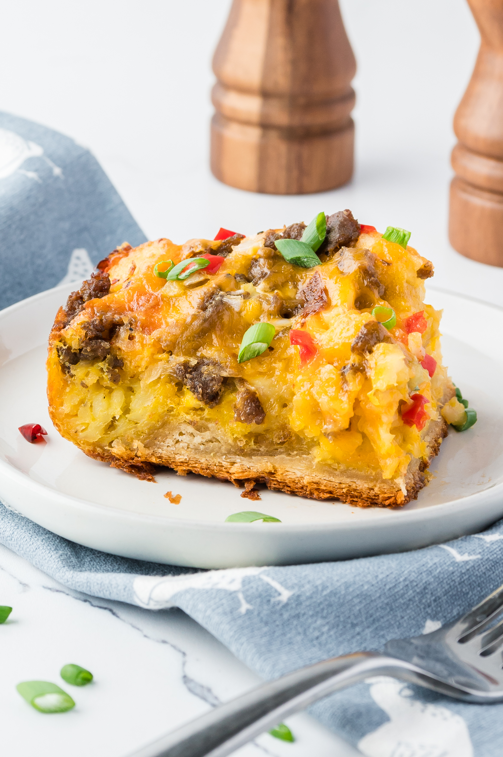 A slice of cheesy breakfast casserole with sausage and vegetables on a white plate, garnished with green onions. Two pepper mills are in the background.