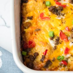 Close-up of a baked dish with melted cheese, ground meat, red bell peppers, and green onions in a white rectangular casserole dish, resting on a blue cloth with a white pattern.