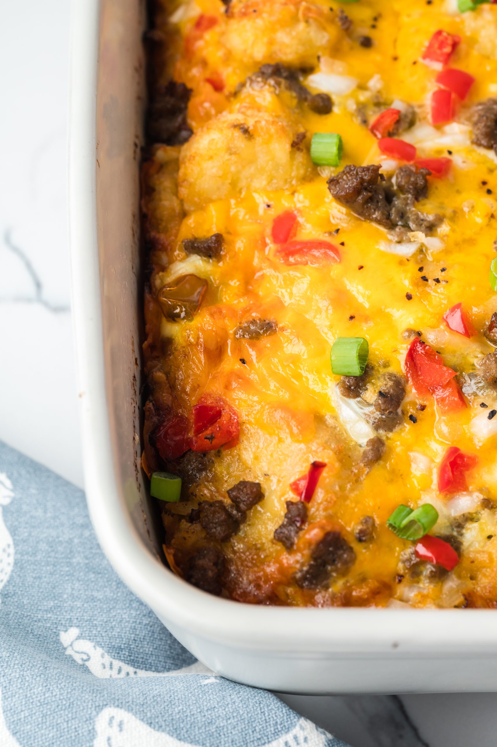 Close-up of a baked dish with melted cheese, ground meat, red bell peppers, and green onions in a white rectangular casserole dish, resting on a blue cloth with a white pattern.