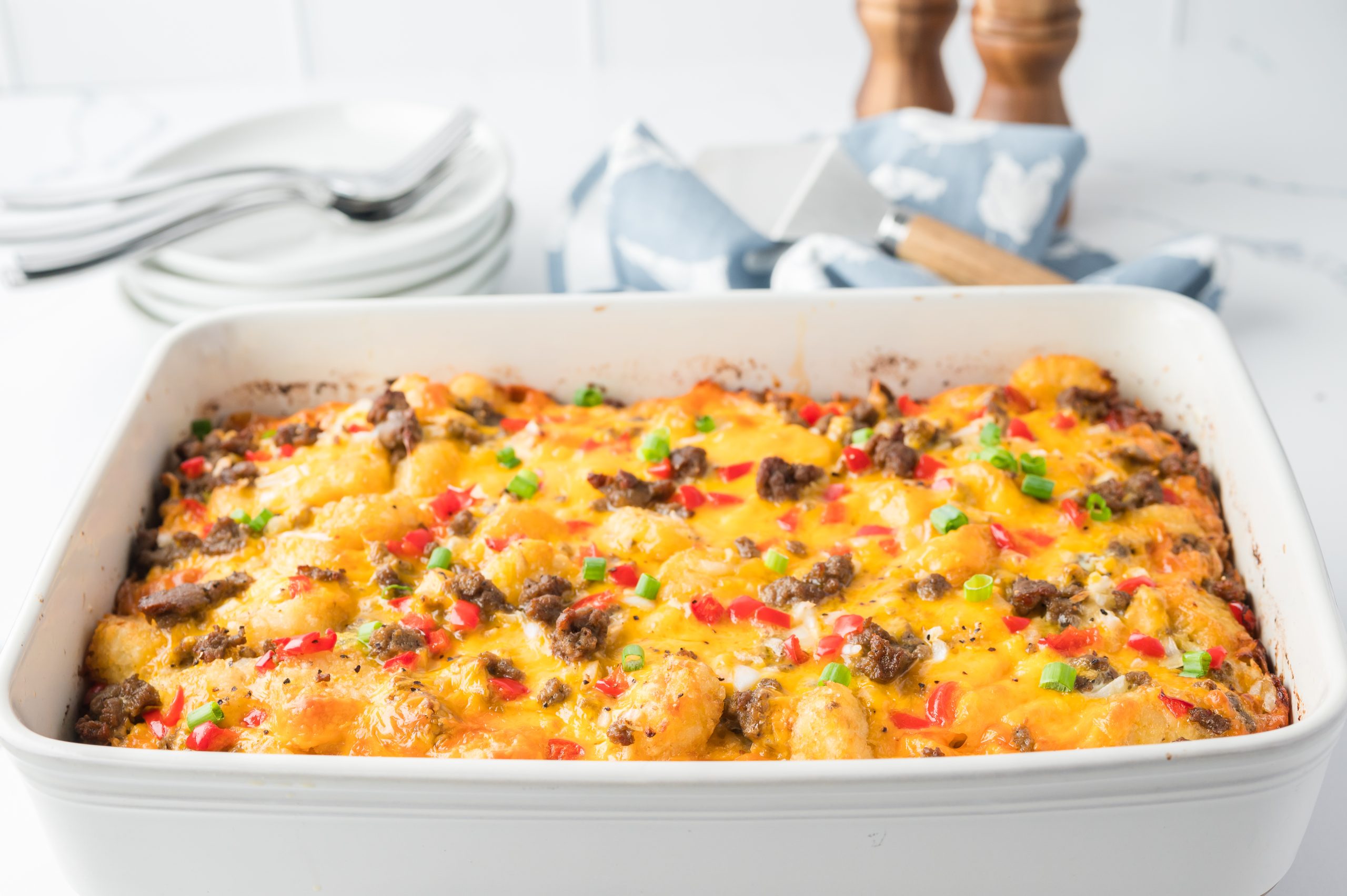 A baked casserole topped with melted cheese, ground meat, red peppers, and green onions in a white dish. Plates and utensils are in the background.