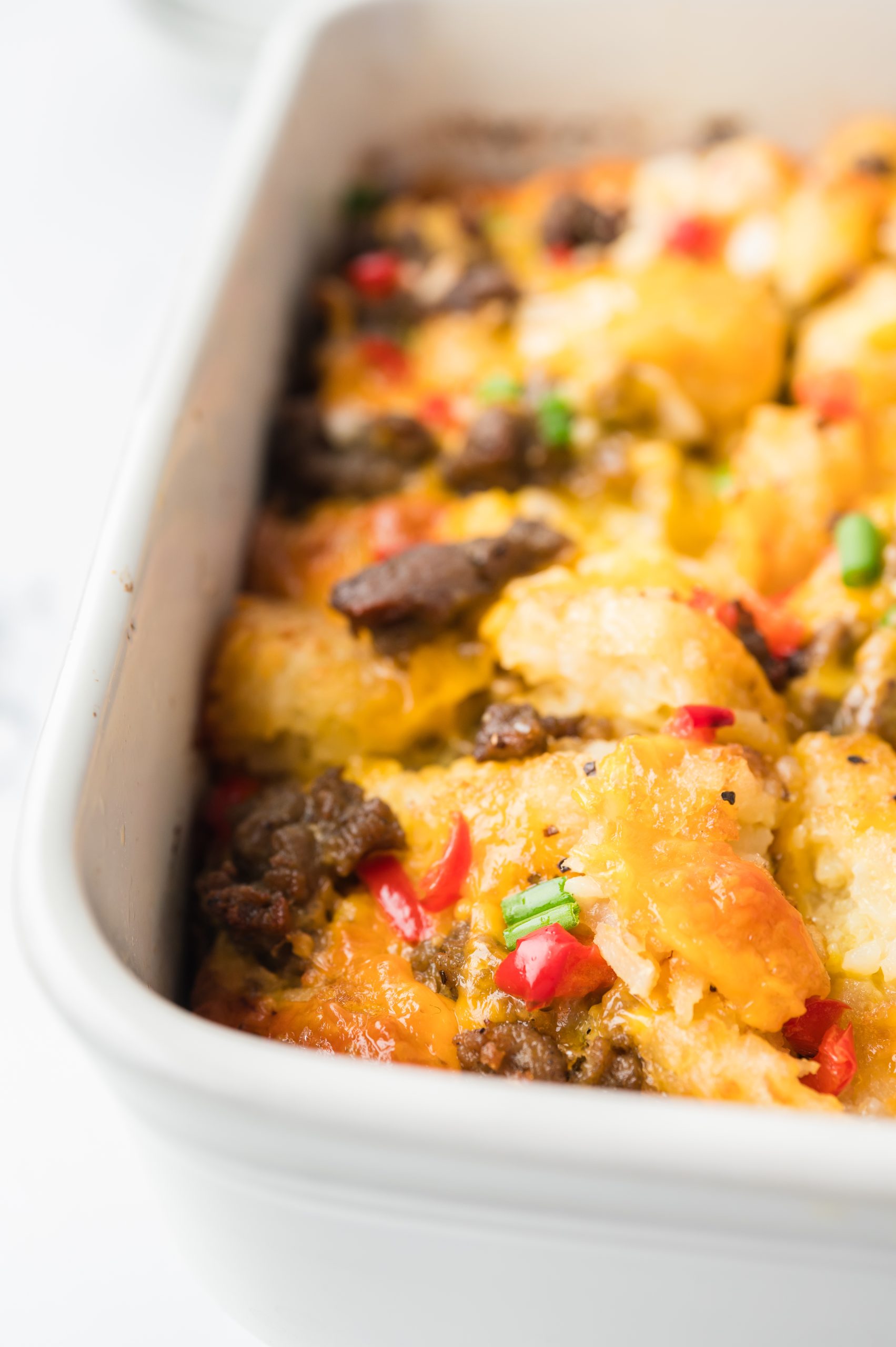 A baked casserole in a white dish, featuring golden bread cubes, browned meat, melted cheese, and garnished with red and green chopped vegetables.