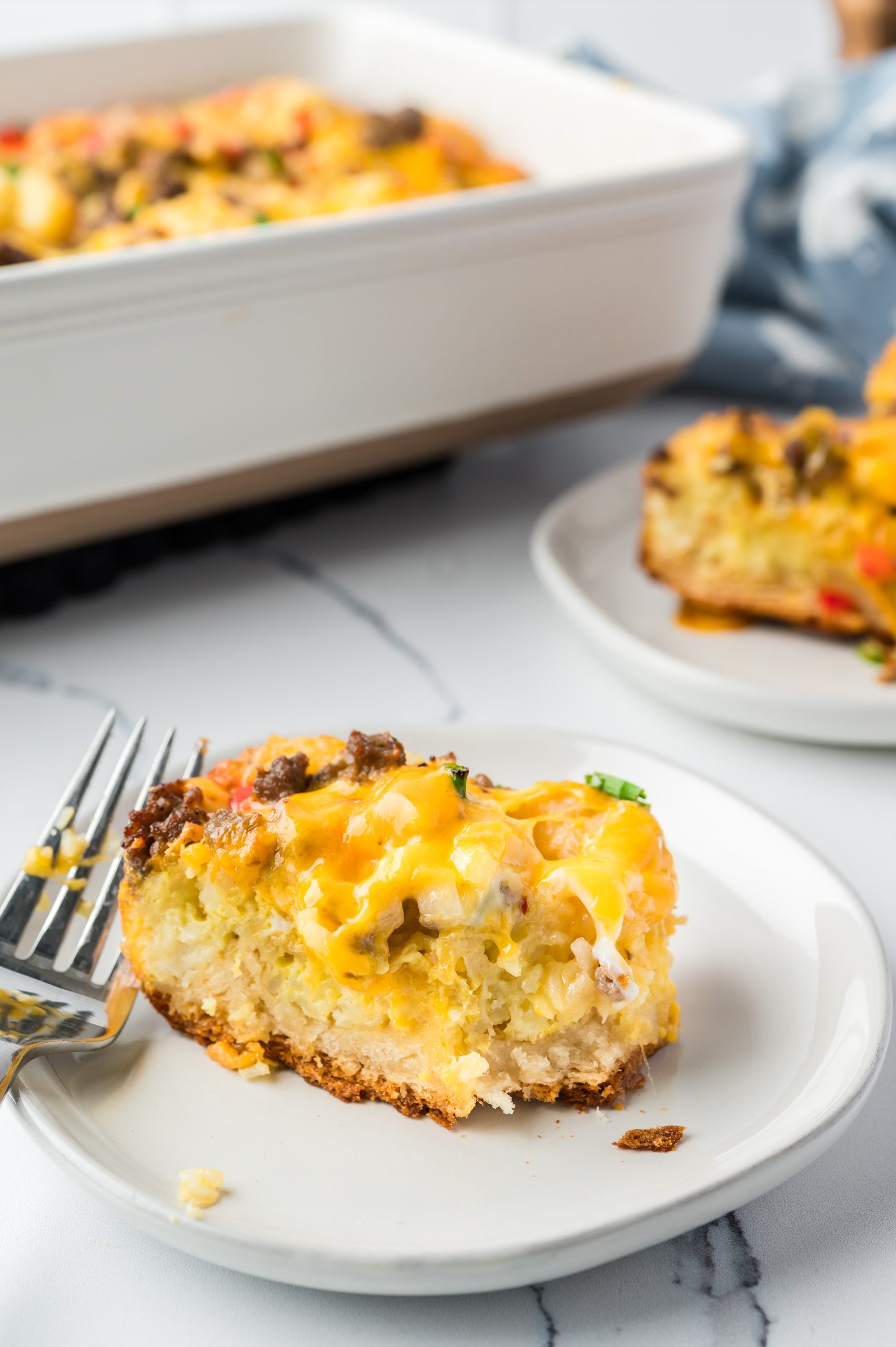 A slice of breakfast casserole with cheese and vegetables on a white plate, with a fork beside it. A larger casserole dish is in the background.