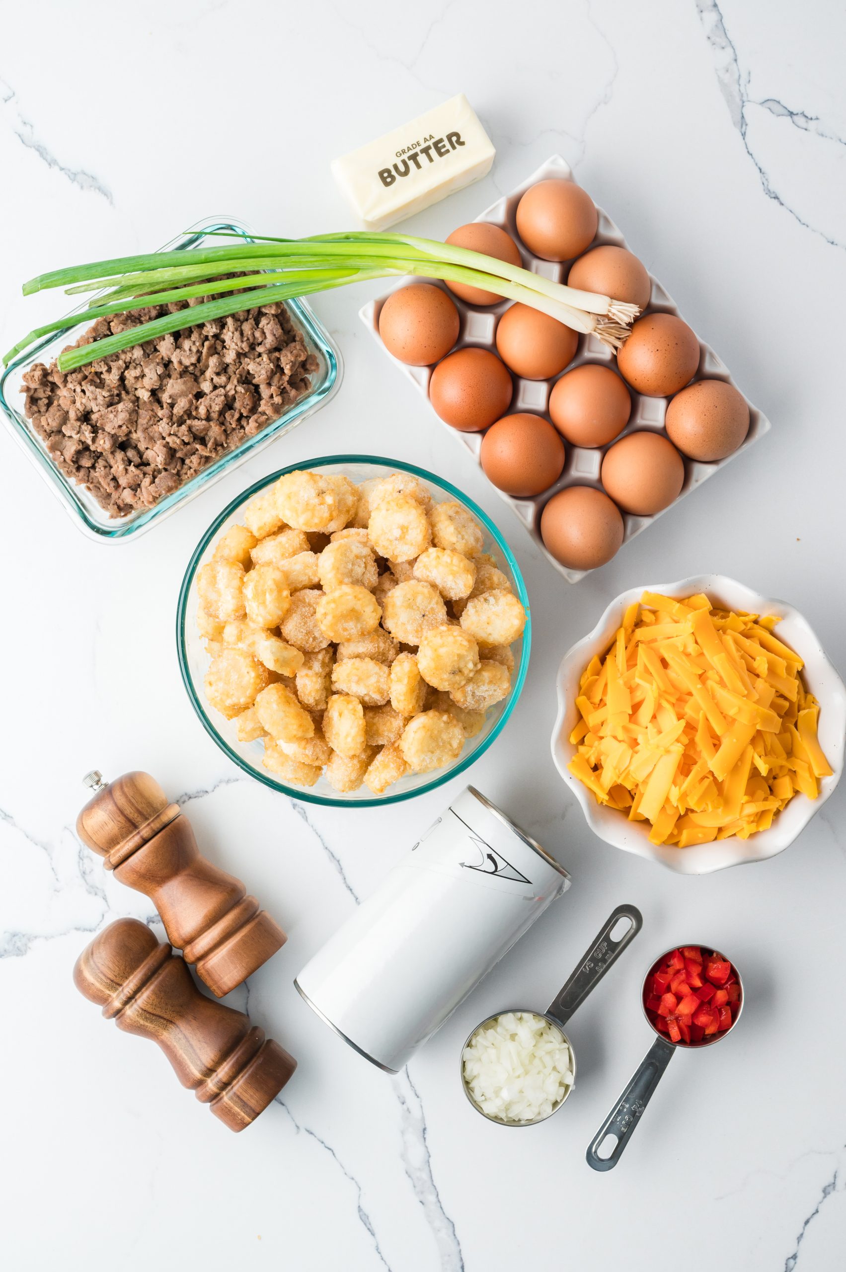 Ingredients on a marble counter: ground beef, eggs, scallions, butter, tater tots, shredded cheese, salt, pepper, chopped onions, and diced red peppers.